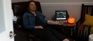 A woman relaxes in a chair with medical sensors on her arm, near a monitoring device and a glowing salt lamp.