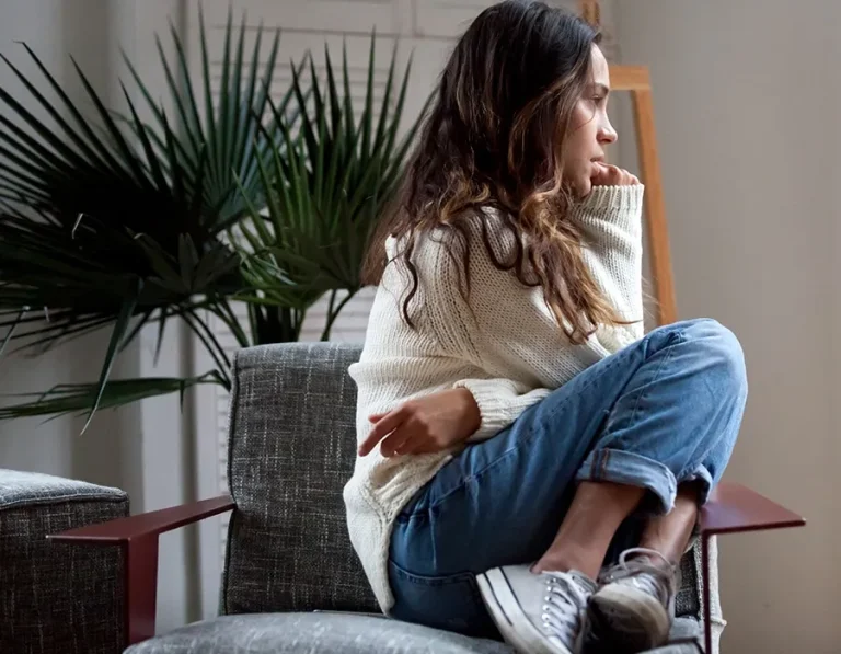 A woman in a sweater and jeans sits curled up on a chair, looking contemplative.