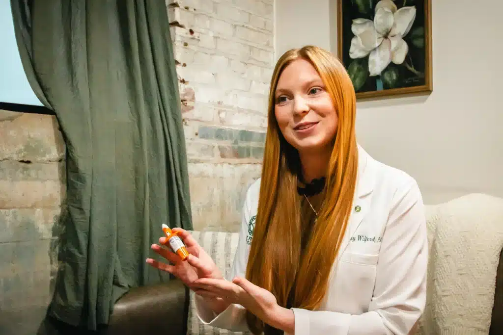 A woman in a lab coat holds a small bottle, gesturing with it.