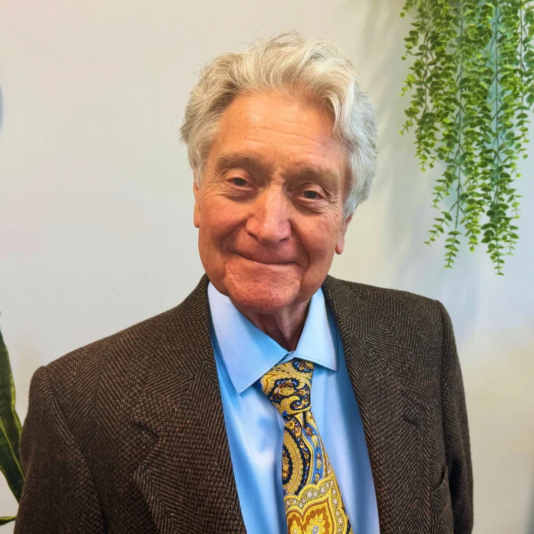 Elderly man in a brown suit with a colorful tie, smiling in front of a white background with hanging plants.