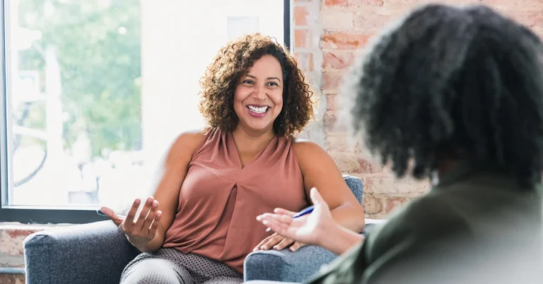 Two people are seated and talking, one smiling brightly, in a casual setting.