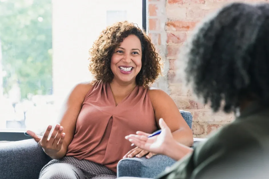 A woman is smiling and gesturing while talking to another person in a casual setting.