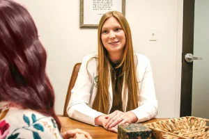 A woman in a white coat smiles while speaking with another person across a table.