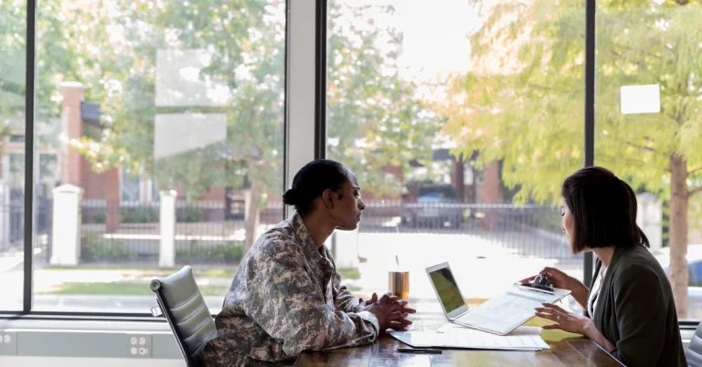 A person in military uniform and another person are having a meeting at a table with a laptop.