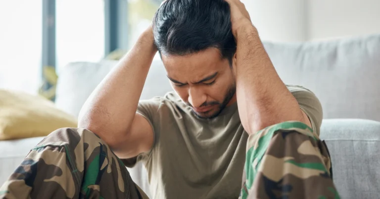Man in camouflage pants sitting with his hands on his head, looking distressed.