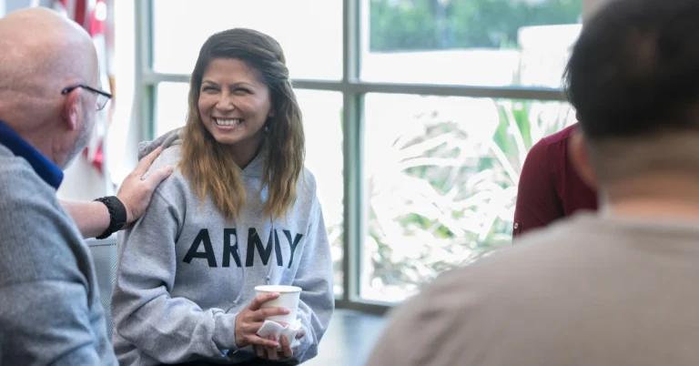 A smiling woman in an "ARMY" hoodie holds a cup while talking with others in a bright room.