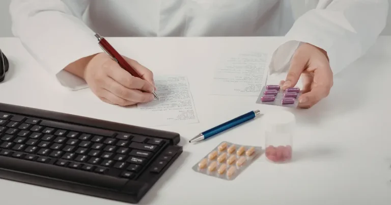 A doctor in a white lab coat writing a prescription while reviewing medication capsules and blister packs on a desk during a medication assisted treatment consultation