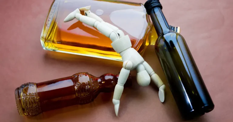 A wooden mannequin figure lying helplessly among alcohol bottles symbolizing the grip of alcohol use disorder and the need for addiction treatment
